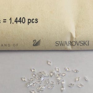 Op de afbeelding: Een verzameling heldere, marquise-vormige kristallen verspreid op een witte ondergrond. De kristallen zijn klein en facetgeslepen, waardoor ze licht reflecteren. Een papieren envelop met de tekst "= 1.440 pcs" en het Swarovski-logo is zichtbaar.