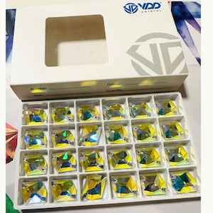 Op de afbeelding: Een transparante plastic container met 20 vierkante, iriserende strass steentjes. De strass steentjes zijn gerangschikt in vier rijen van vijf. De container is gelabeld met "VD Crystal".