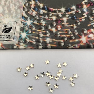 May include: Small, gold-coloured star-shaped embellishments scattered on a white surface. The word "PRECIOSA" is visible on a partially visible package in the background, along with a "lead-free brilliance" logo.