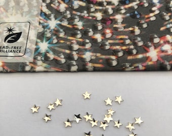 May include: A collection of small, gold-colored star-shaped embellishments scattered on a white surface. The word "PRECIOSA" is visible on a dark background, along with a logo indicating lead-free brilliance.