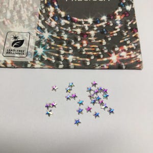 May include: A collection of small, iridescent star-shaped crystals scattered below a Preciosa brand packaging card. The card features the text 438 11 612, PRECIOSA, and a logo with the text LEAD-FREE BRILLIANCE, alongside a graphic of circular crystals.
