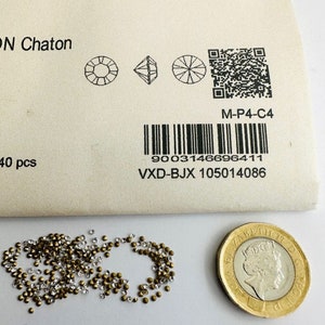 Könnte beinhalten: Nahaufnahme einer kleinen Tüte mit 40 klaren und goldenen Strasssteinen. Die Tüte ist mit dem Markennamen "Swarovski" und dem Produktcode "M-P4-C4" gekennzeichnet.