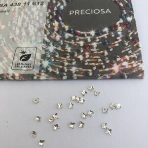 Könnte beinhalten: Kleine, silberfarbene, schmetterlingsförmige Strasssteine, die auf einer weißen Oberfläche verstreut sind. Im Hintergrund befindet sich eine Karte mit dem Wort "PRECIOSA" und dem Text "LEAD-FREE BRILLIANCE".