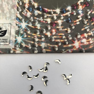 May include: Silver crescent-shaped embellishments scattered on a white surface. The embellishments are reflective and appear to be made of metal. The top of the image shows a package with the word "PRECIOSA" printed on it.