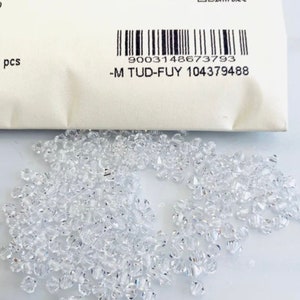 May include: A close-up of a pile of clear glass beads. The beads are small and have a faceted design. The beads are packaged in a white bag with a barcode and the text -M TUD-FUY 104379488.