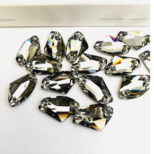 Puede incluir: Un grupo de piedras de cristal transparentes y facetadas en forma de lágrima con dos agujeros en la parte superior. Las piedras están esparcidas sobre una superficie blanca.