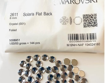 May include: A package of 144 clear, foiled, 8 mm Swarovski Solaris flat back crystals. The package is labeled with the product number 2611, the size, and the Swarovski logo. The crystals are scattered on a white surface.