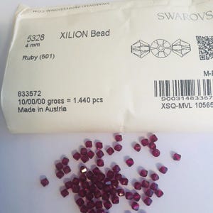 Könnte beinhalten: Eine Ansammlung von rubinroten XILION-Perlen, 4 mm groß, auf einer weißen Oberfläche. Die facettierten Perlen reflektieren das Licht. Ein Papierpaket mit dem Text "XILION Bead" und "Ruby (501)" befindet sich im Hintergrund.