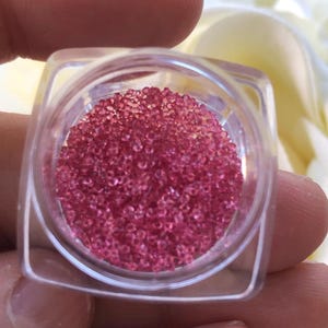 Puede incluir: Un pequeño recipiente transparente lleno de microperlas rosas. El recipiente está en una mano.