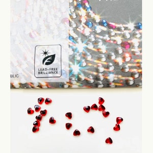 Op de afbeelding: Een strooiing van rode hartvormige strasssteentjes op een wit oppervlak. De strasssteentjes zijn klein en hebben een heldere, glanzende afwerking.