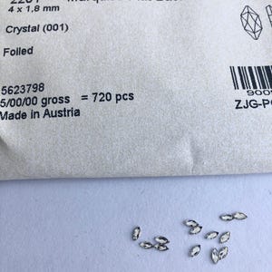 以下が含まれることがあります： 「Marquise Flat Back」とラベルされたパッケージのクローズアップ。サイズは4 x 1.8 mm。パッケージには、小さなクリスタルのようなマーキス型の装飾品が入っています。「Made in Austria」の文字が見えます。