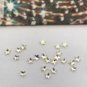 May include: A scattering of small, gold-coloured star-shaped embellishments on a white surface. The stars are reflective and catch the light. A patterned fabric is visible in the background.