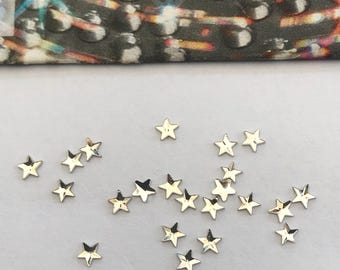 May include: A collection of small, metallic gold star-shaped embellishments scattered on a white surface. The stars vary slightly in size and are reflective, catching the light. A patterned fabric is visible in the background.