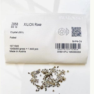 May include: A scattering of small, round crystals next to a white packet. The packet, branded "SWAROVSKI," details "XILION Rose" crystals, size SS 10, and other information. The crystals are tiny, with a reflective, metallic appearance.