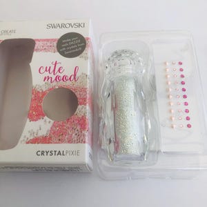 Puede incluir: Un juego Swarovski Crystal Pixie en un recipiente de plástico transparente. El juego incluye una pequeña botella de purpurina, un paquete de pequeños cristales y una caja con las palabras "cute mood" y "CRYSTALPIXIE".
