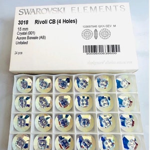 Könnte beinhalten: Eine Schachtel mit 24 klaren, irisierenden 18 mm Rivoli CB (4 Löcher) Kristallen von Swarovski Elements. Die Kristalle sind in einem Raster in einer weißen Schachtel angeordnet. Die Schachtel trägt die Produktinformationen, einschließlich der Produktnummer, Größe, Farbe und Menge.