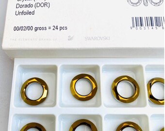 May include: A tray of gold-colored, faceted crystal rings, each 20 mm in diameter, labeled "Cosmic Ring." The rings are arranged in a white tray with individual compartments. The packaging includes the Swarovski logo and product information.