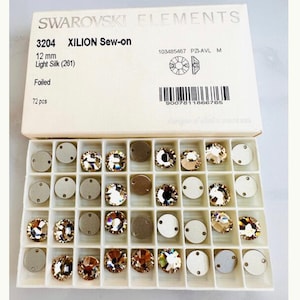 May include: A clear plastic container with 72 small, iridescent, gold-toned sew-on crystals. The container is labelled "Swarovski Elements 3204 XILION Sew-on 12 mm Light Silk (261) Foiled 72 pcs".