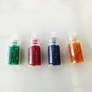 May include: Four small glass bottles filled with colorful beads. The bottles are clear and have a small opening at the top. The beads are green, red, blue, and orange.