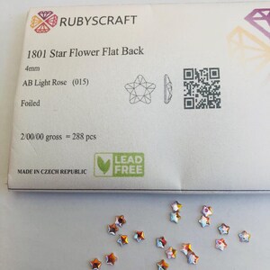 以下が含まれることがあります： 「1801 Star Flower Flat Back」と「AB Light Rose (015)」とラベルされた星型のフラットバックラインストーンのパッケージ。 ラインストーンは約4mmで、明るい表面に散らばっています。 パッケージには「LEAD FREE」の文字も含まれています。