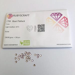 Op de afbeelding: Een verpakking hartvormige flatback strass steentjes in licht amethist kleur, 3 mm groot. De verpakking bevat de tekst "RUBYSCRAFT", "1789 Heart Flatback" en "MADE IN CZECH REPUBLIC". Een aantal steentjes liggen verspreid.