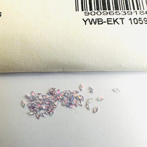 Pode incluir: Uma dispersão de strass iridescentes, transparentes, facetados, em forma de diamante sobre um fundo branco.