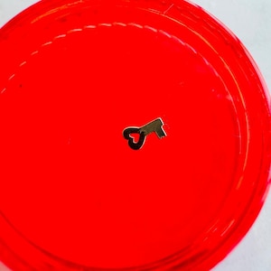 Puede incluir: Un dije de llave de color dorado con un lazo en forma de corazón en la parte superior, sentado sobre una superficie de color rojo brillante.