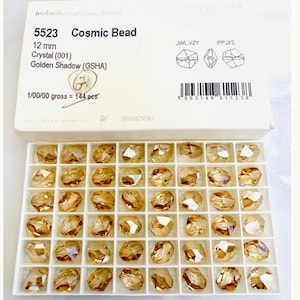 Może przedstawiać: Pudełko z 144 złotymi kryształami Swarovski Cosmic Bead, każdy o wymiarach 12 mm. Pudełko jest oznaczone numerem produktu 5523, kolorem kryształu "Golden Shadow" i rozmiarem "12 mm".