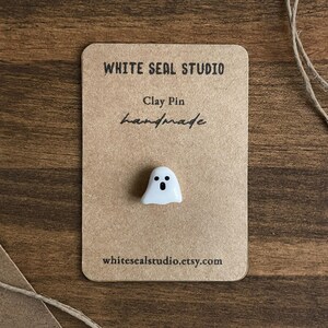 May include: A handmade white clay ghost pin with black eyes and mouth, displayed on a brown card. The card features the text "WHITE SEAL STUDIO", "Clay Pin", "handmade", and an Etsy shop URL.