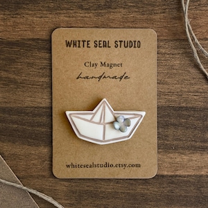 Puede incluir: Un imán de arcilla hecho a mano con forma de barco de papel, con una pequeña flor. El imán es blanco con un contorno marrón claro y una pequeña flor azul-gris. Se presenta en una tarjeta marrón con el texto "WHITE SEAL STUDIO", "Clay Magnet" y "handmade".