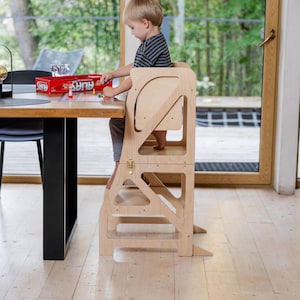 Puede incluir: Una silla de madera para niños con un escalón integrado. La silla es de color marrón claro y tiene un diseño sencillo. Un niño está sentado en la silla y jugando con juguetes en una mesa.