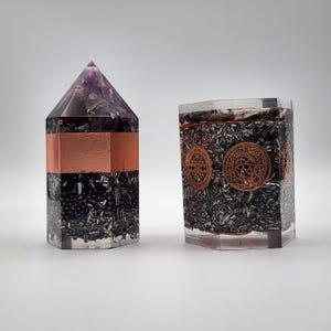 May include: Two orgone energy devices. One is a tall, hexagonal pyramid with a copper band and a purple crystal top. The other is a shorter, hexagonal shape with copper-colored circular designs.
