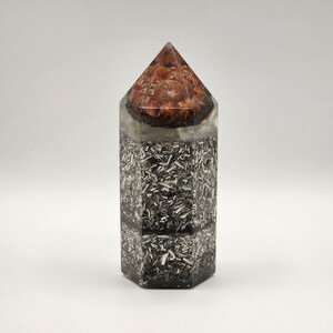 May include: A hexagonal orgone pyramid with a clear resin exterior. The top section features a cluster of reddish-brown stones, while the lower section is filled with silver-colored metal shavings. The pyramid has a pointed top.