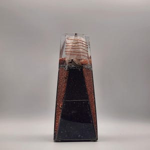 Può includere: Una piramide di resina rettangolare trasparente con un design a strati. La piramide presenta una base nera, lati color rame e una sezione superiore trasparente con una bobina di rame e pietre. È un oggetto decorativo.