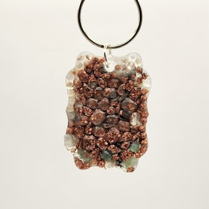 May include: A copper and green glitter resin keychain with a silver key ring. The keychain is shaped like a rectangle with rounded corners.