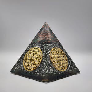 May include: A black and silver pyramid-shaped orgone energy generator with two gold-colored flower of life symbols. The top of the pyramid features a copper coil and a green and brown design.
