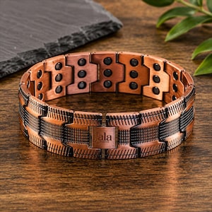 Könnte beinhalten: Kupferfarbenes Armband mit schwarzen Akzenten und einem rechteckigen Verschluss. Das Armband hat eine Reihe von rechteckigen Gliedern mit eingebetteten schwarzen Steinen. Der Verschluss ist mit dem Wort "Jala" und dem Wort "studios" graviert. Das Armband liegt auf einer Holzoberfläche.