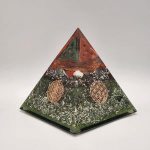 May include: A handcrafted orgone pyramid with a green base, featuring copper, metal shavings, and decorative elements. The pyramid has a layered design with a mix of colors and textures, including a copper wire coil and two golden flower of life symbols.