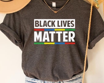May include: Dark grey heather V-neck t-shirt with the text "Black Lives Matter" in white with a rainbow stripe underneath. The shirt is hanging on a wooden hanger with a brown hat and a woven bag.
