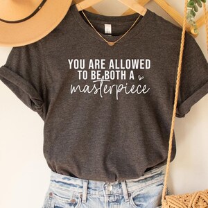 May include: Dark grey heather t-shirt with the text "YOU ARE ALLOWED TO BE BOTH A masterpiece" in white. The shirt is paired with denim shorts, a tan hat, and a woven crossbody bag.