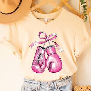 May include: A cream colored t-shirt with a pink ribbon tied around a pair of pink boxing gloves. The shirt is hanging on a wooden hanger and has a soft cream color.