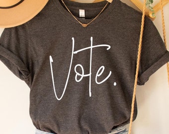 May include: Dark grey heather V-neck t-shirt with the word "Vote" written in white cursive script. The shirt is hanging on a wooden hanger with a straw hat and a woven bag.