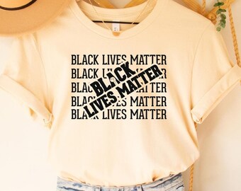 May include: A soft cream colored t-shirt with the text "Black Lives Matter" repeated multiple times. The text is crossed out with a diagonal line. The shirt is on a hanger and is being modeled on a white background.