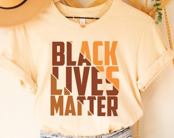 May include: A cream colored t-shirt with the text "BLACK LIVES MATTER" printed in brown and orange. The shirt is hanging on a wooden hanger and is styled with denim shorts and a straw bag.