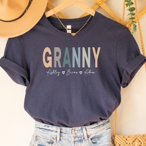 May include: Navy blue t-shirt with the word "Granny" in colorful letters and the names "Ashley, Brian, & Aiden" written below. The shirt is hanging on a wooden hanger with a straw hat and a small woven bag.