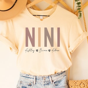 May include: A soft cream colored t-shirt with the word "NINI" printed in large letters in shades of gray and brown. The text "Ashley ♥ Brian ♥ Adam" is printed below the word "NINI".