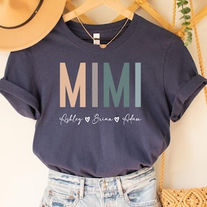May include: A navy blue t-shirt with the word "MIMI" in colorful block letters and the names "Ashley", "Brian", and "Eden" written below. The shirt is hanging on a wooden hanger with a straw hat and a woven bag.