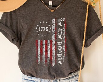 May include: Dark grey heather V-neck t-shirt with a distressed American flag design and the text "We The People" and "1776".