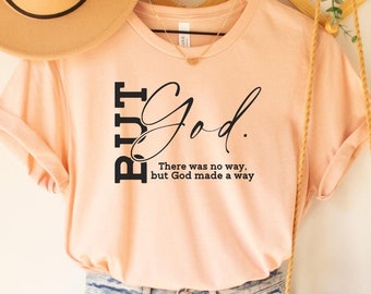 May include: A peach-colored t-shirt with the text "BUT God. There was no way, but God made a way" printed on the front. The shirt is hanging on a wooden hanger with a straw hat and a woven bag.