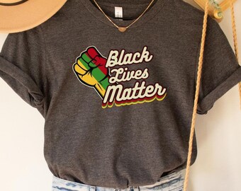 May include: Dark grey heather V-neck t-shirt with a black fist raised in the air and the text "Black Lives Matter" in a retro font. The shirt is hanging on a wooden hanger with a straw hat and a woven bag.